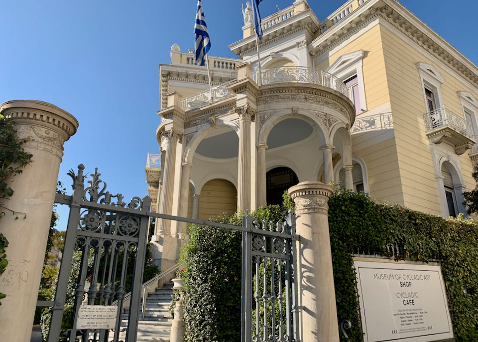Exterior of a yellow neoclassical mansion with white pillars and a Greek flag