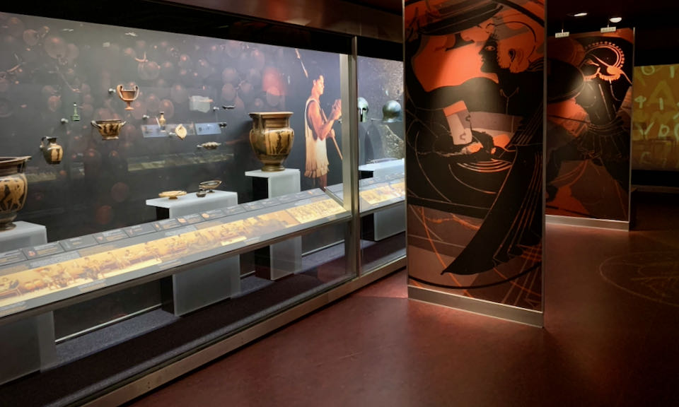 Items on display in a museum case, alongside a mural of ancient Greek life