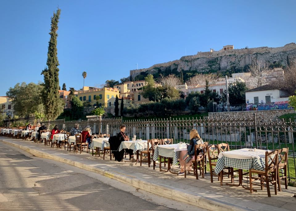 Diners sit at outdoor tables on a sidewalk with views of the Acropolis.