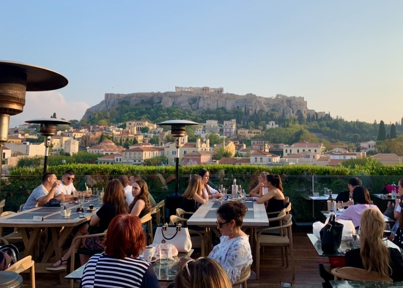 People dine at outdoor tables with a sunset view of the Acropolis in Athens