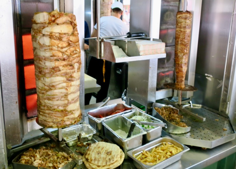 Window of a gyro joint, with meat on a spit, stacked pitas, and containers of condiments