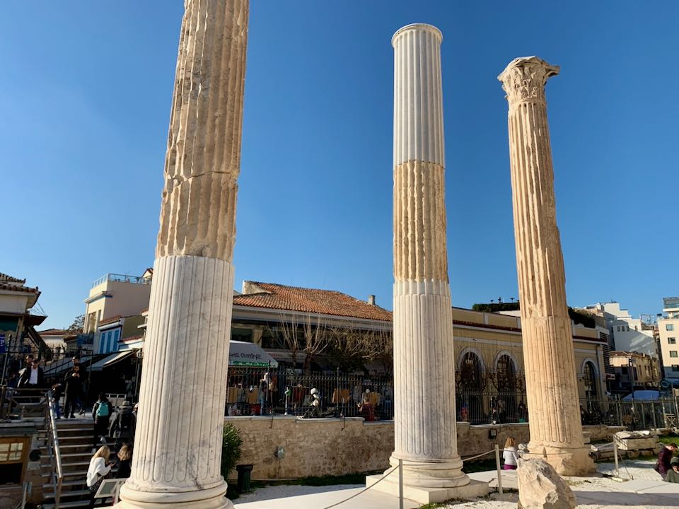 View through three reconstructed ancient columns to the modern street above, crowded with cars and pedestrians.