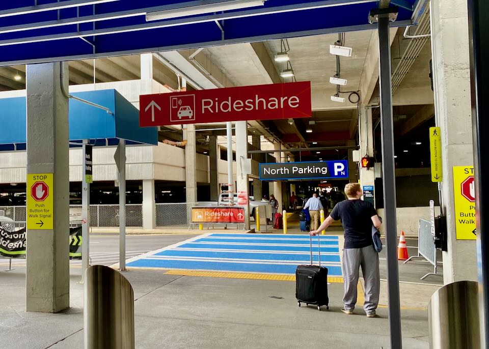 Following a Rideshare sign, passengers walk toward a blue crosswalk leading to a parking garage.
