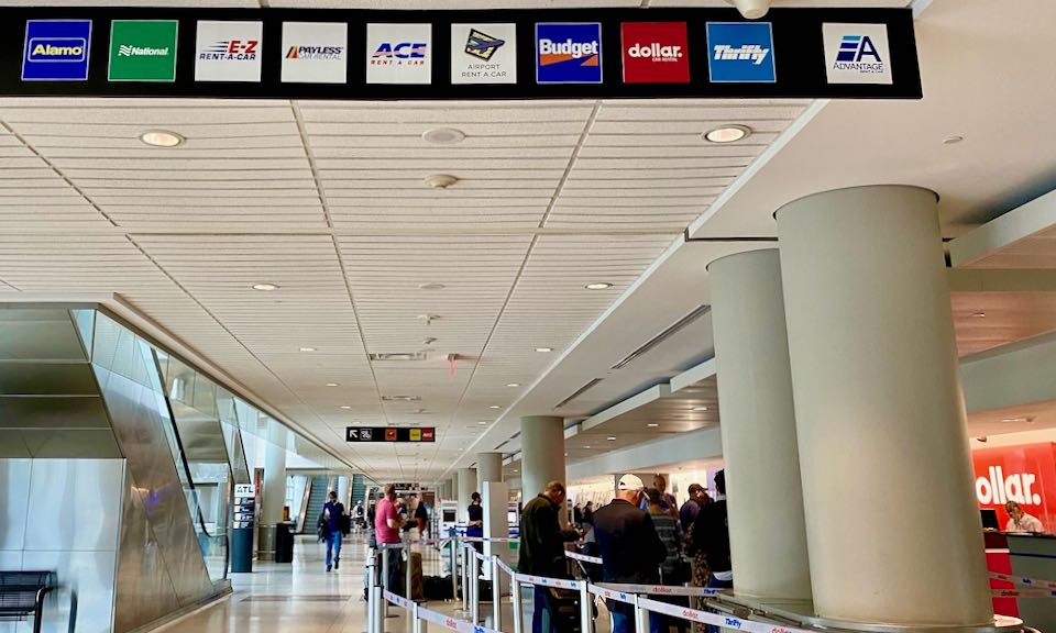 People wait in line at an airport under a sign showing the names of car rental companies