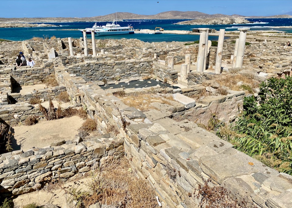 View across a landscape of ancient stone ruins to a modern harbor with a large boat at dock