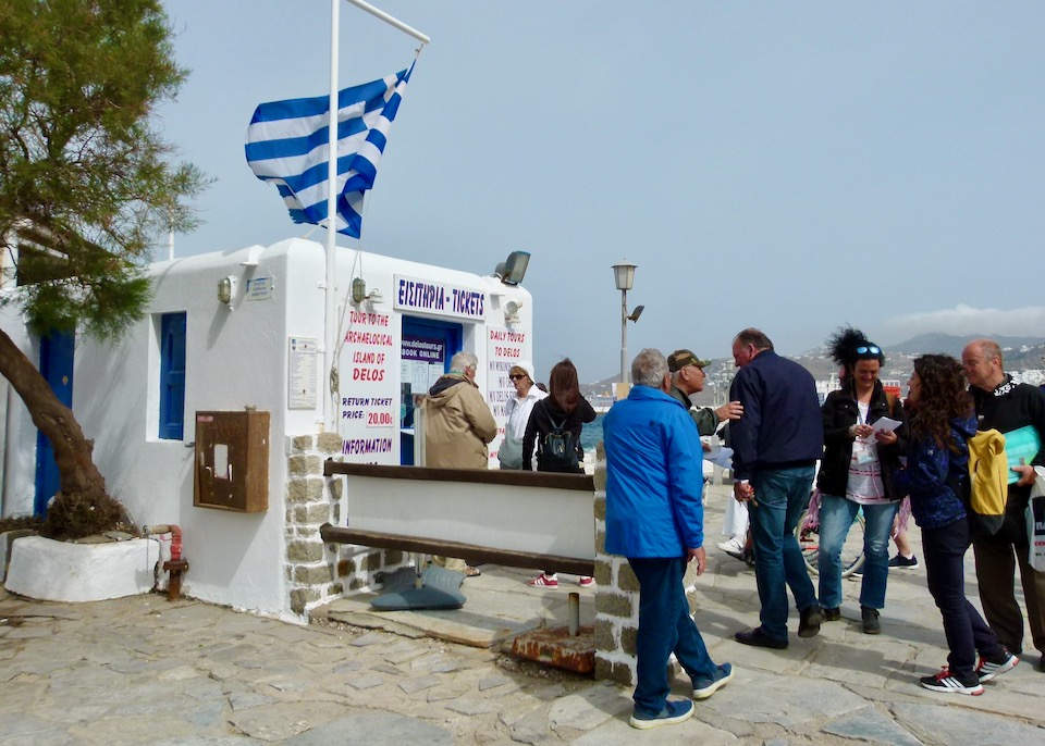 People line up on a windy day at a kiosk selling boat tickets to Delos Island. A Greek flag flaps wildly.