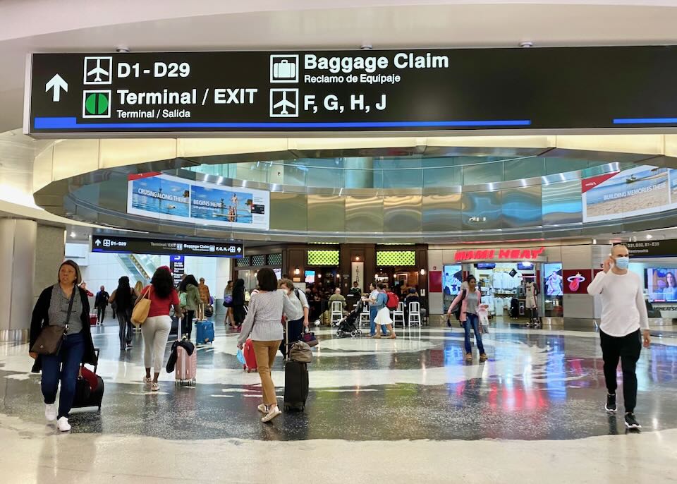 An overhead sign in the airport directs passengers to the exit, baggage claim, and D, F, G, H, and J terminals.
