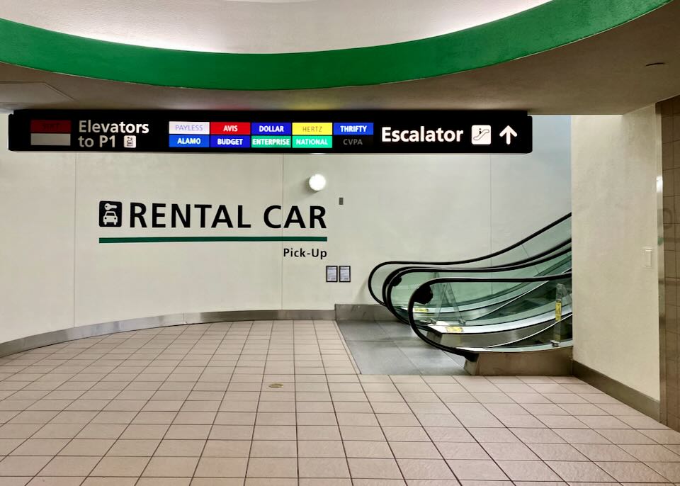 A sign shows that by taking the escalator up, there are Payless, Avis, Dollar, Hertz, Thrifty, Alamo, Budget, Enterprise, and National rental car agencies.