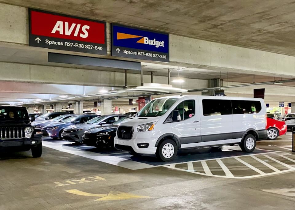 A white Ford passenger van sits in the Avis and Budget parking lot.