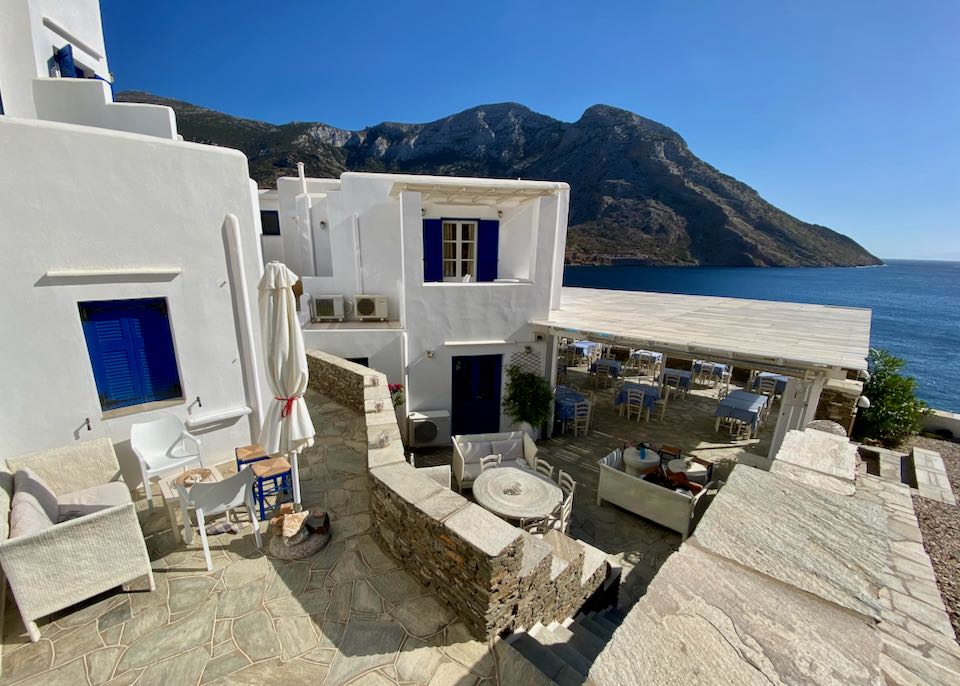 View past the stone terrace of a white, boxy hotel to a crystal blue bay and mountains.