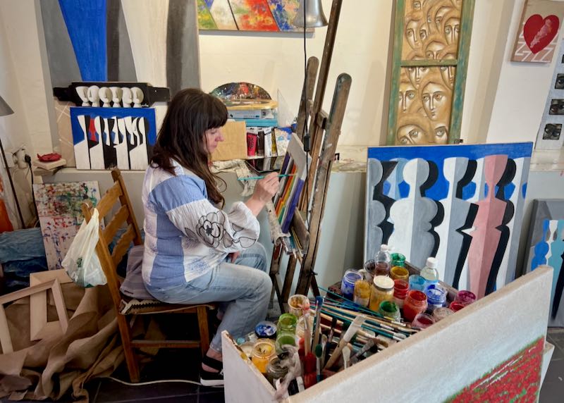 A woman sits at an easel and paints, surrounded by her colorful artwork