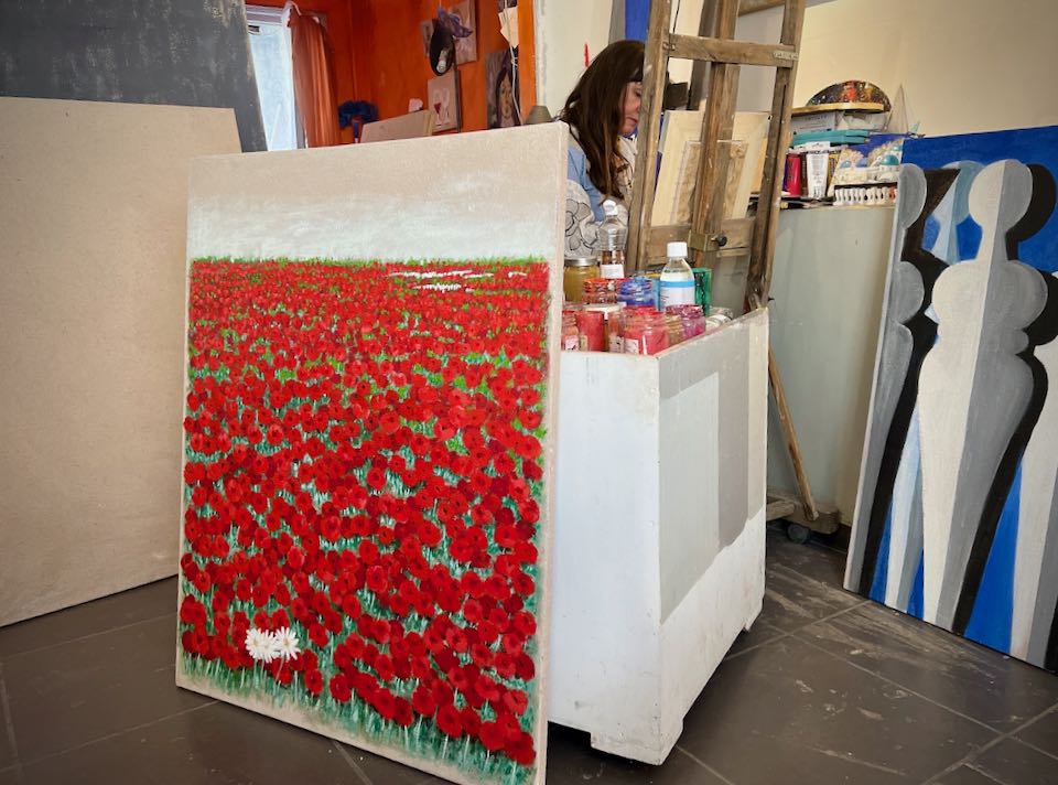 A woman paints at an easel behind a large painting of poppies in a field