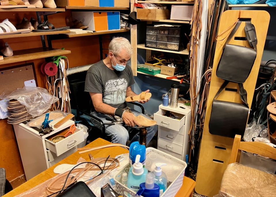 A man sits at a small workbench and crafts a leather sandal.