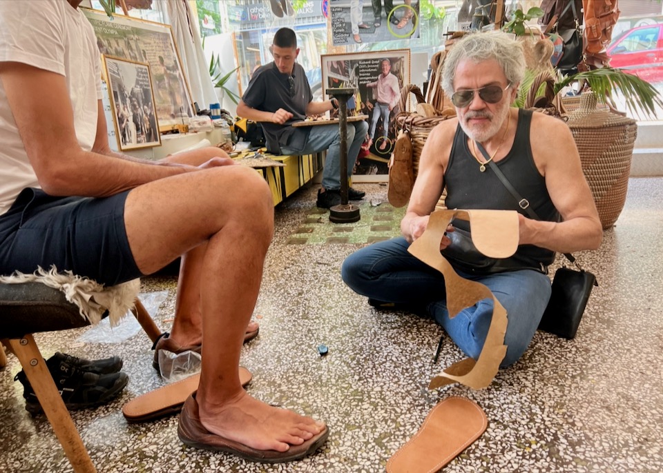 A man sits on the floor and cuts out a template for the sole of a leather sandal.