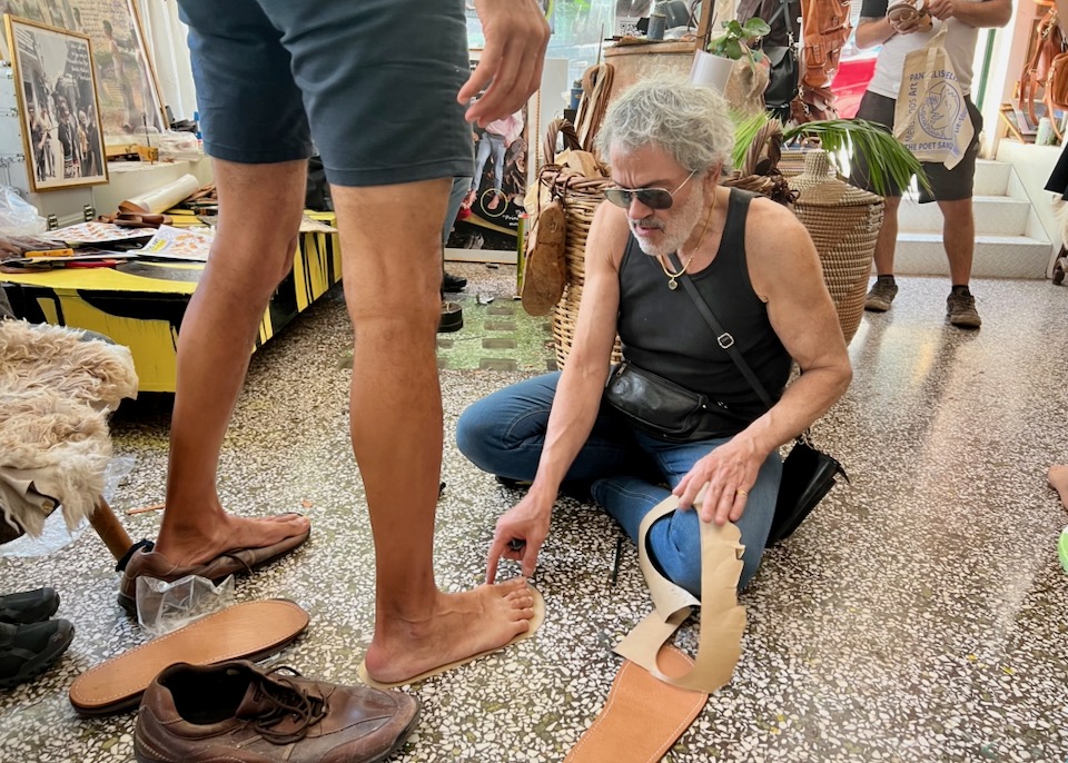 A man sits on the floor and measures another man's foot against a sandal sole template