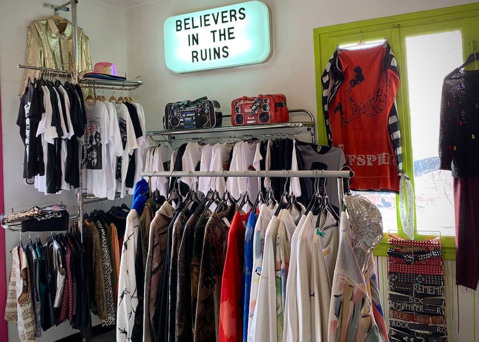 Rack of clothing with a lit sign hanging above that reads "Believers in the Ruins"