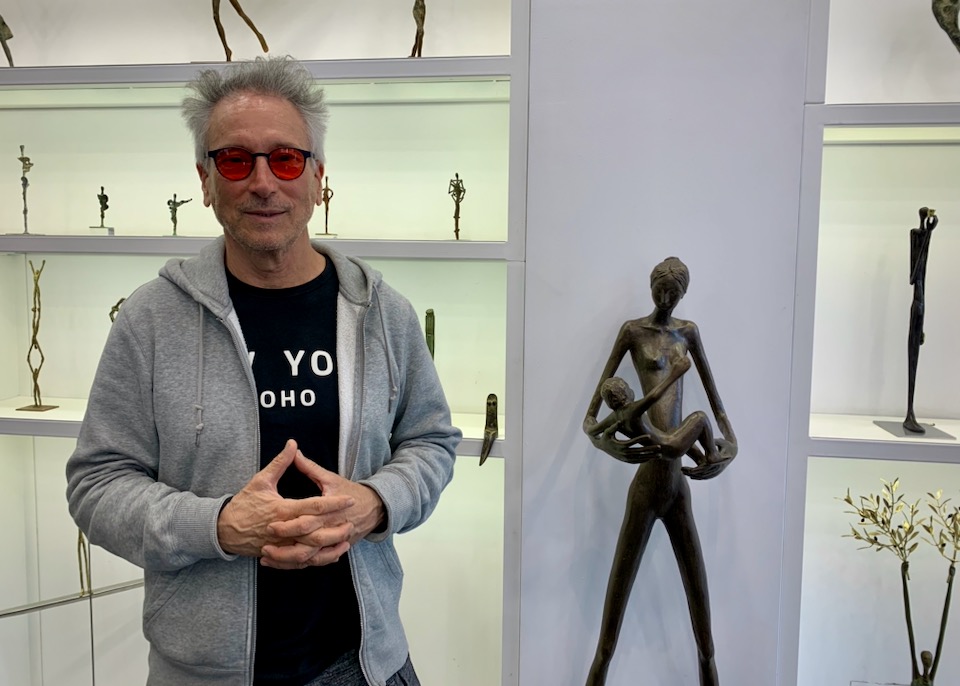 A hip man with grey wavy hair and red eyeglasses stands in front of metal sculptures dislpayed on shelves