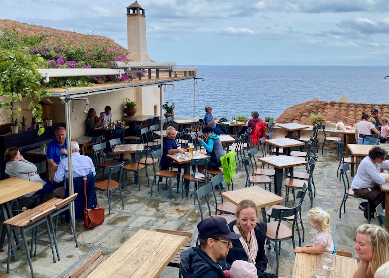 People eat and drink at tables on a stone terrace overlooking the sea