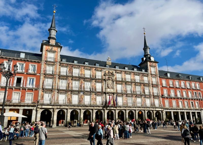 Large ornate buildings lining the Plaza Mayor in Madrid, with many pedestrians walking past.