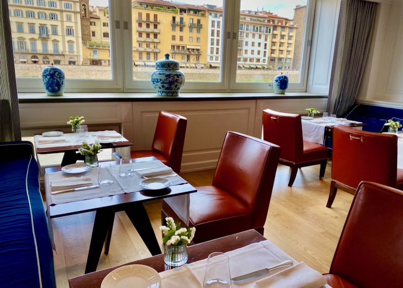 Dining chairs and tables set next to a window overlooking the Arno river 