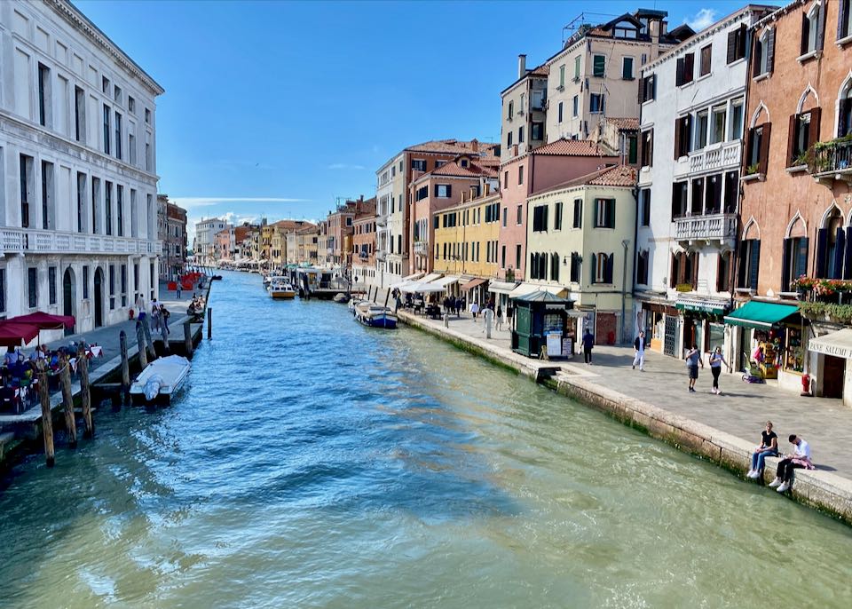Rustic and elegant Venetian buildings line a canal, with two people sitting on its side, enjoying the view