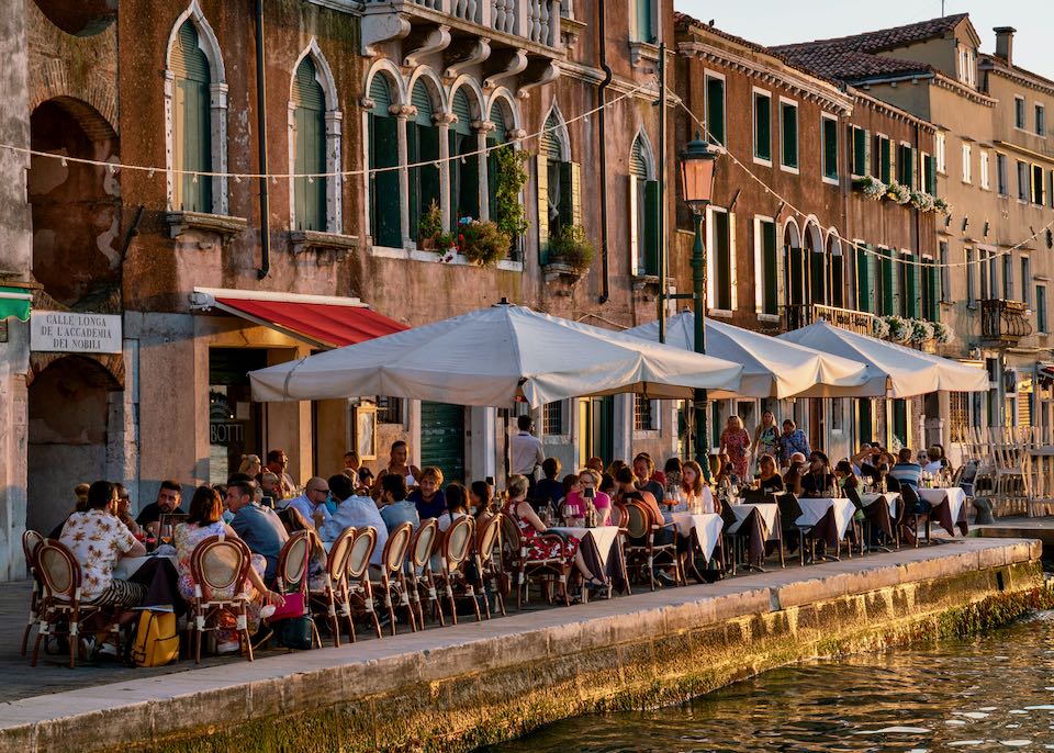 Crowded cafe tables with diners under umbrellas on a Venetian canal at dusk