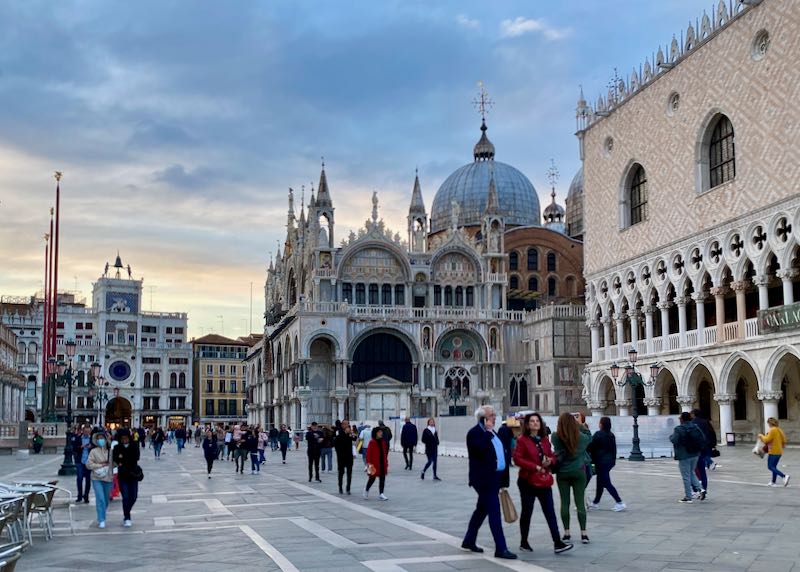 Pedestrians walk past the ornate and colorful St. Marks Basilica and Square in Venice at dusk.