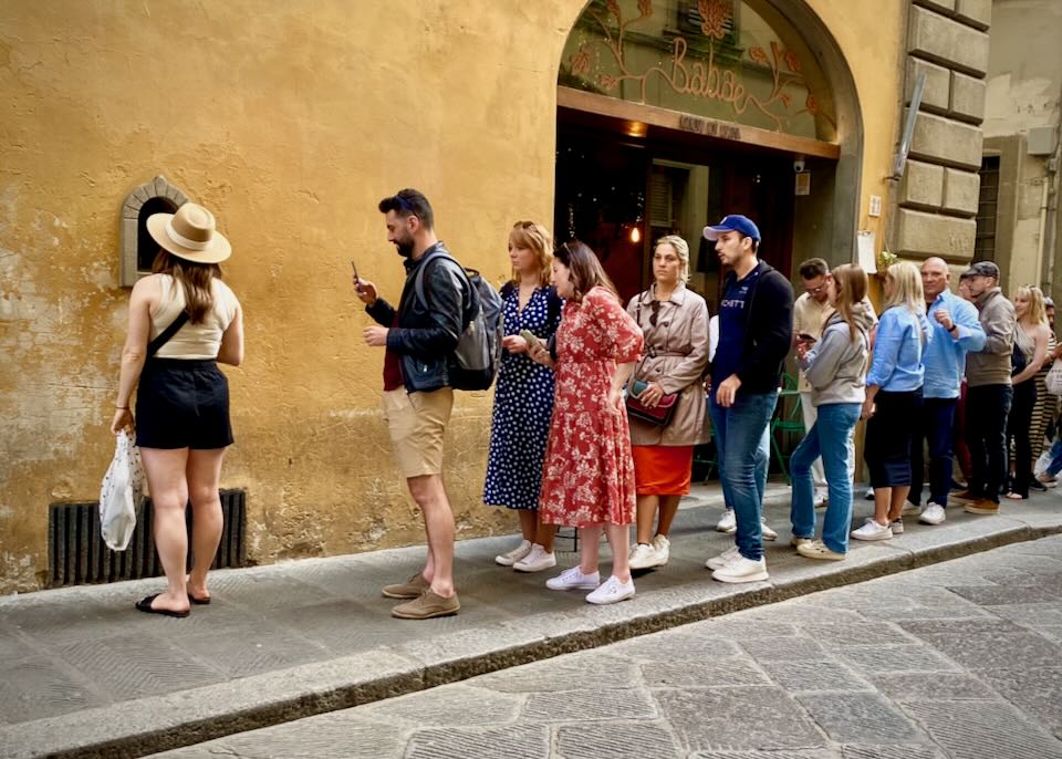 People waiting in line outdoors beside a small window. 