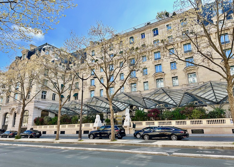 The grand limestone exterior of the Peninsula hotel in Paris with two white Chinese lion statues and a geometric glass and metal awning running the across the full length of the building