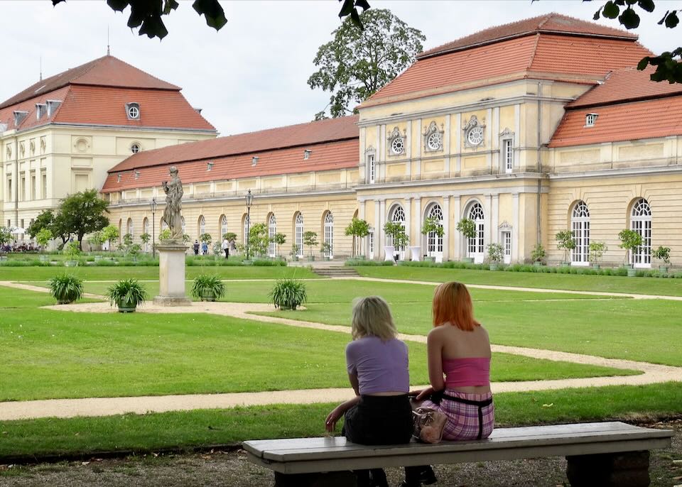 Back view of two teenage girls sitting on a stone bench, facing a green garden courtyard and palace