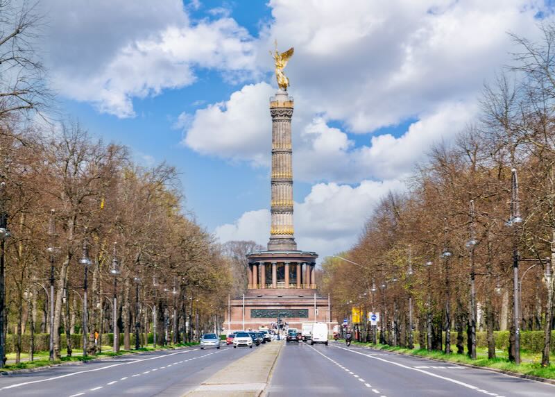 A sunny spring day in the Tiergarten park in the Mitte district of Berlin, Germany. The 17th June Street is in the foreground, and the Victory Column is in the background. The sky is blue with fluffy clouds.