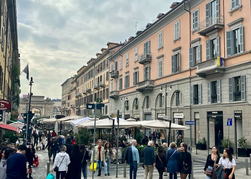 People shop under umbrellas at outdoor stalls on a pedestrianized Italian street