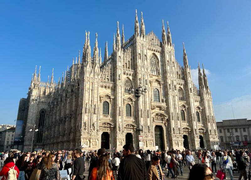 A magnificent, gothic-spired church on a sunny day, with a crowd at the piazza in front