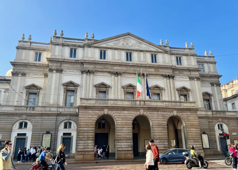 Pedestrians walk past a historic building with an Italian flag flying above the entrance