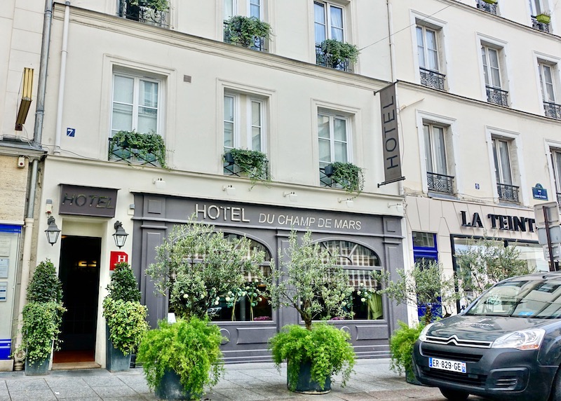 Exterior of the Hotel du Champ de Mars in gray and cream with green vines and potted trees on the balconies and sidewalk in the 7th Arrondissement of Paris
