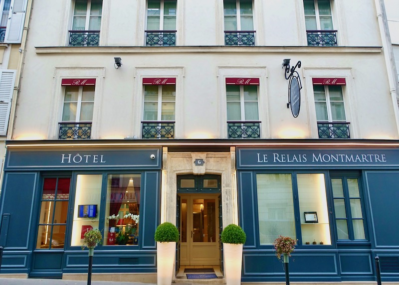Exterior of the Hotel Le Relais Montmartre in blue and cream with red accents, wrought iron railings, and plants in front in the 18th Arrondissement of Paris