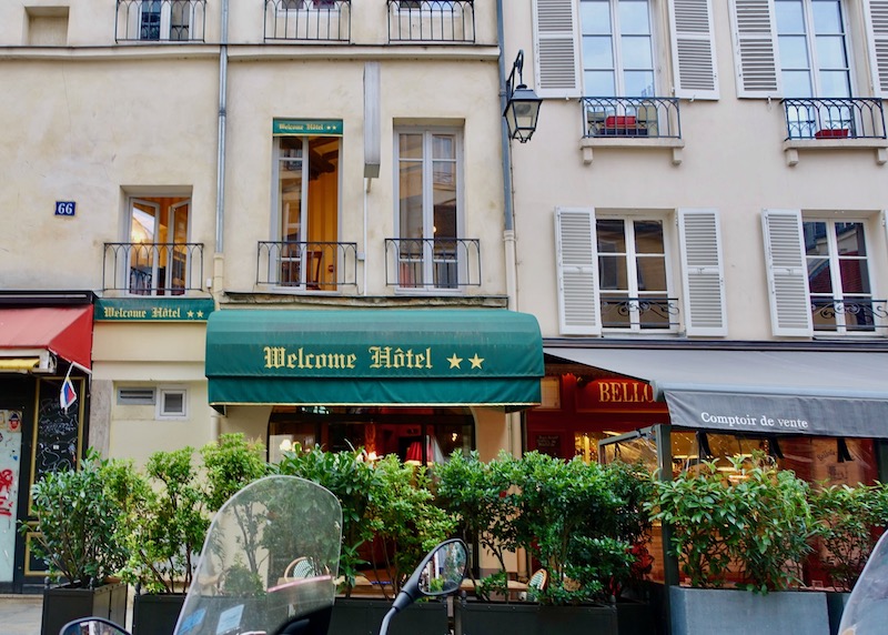 The entrance of the Welcome Hotel with a green and gold awning and a small privacy hedge in the 6th Arrondissement of Paris