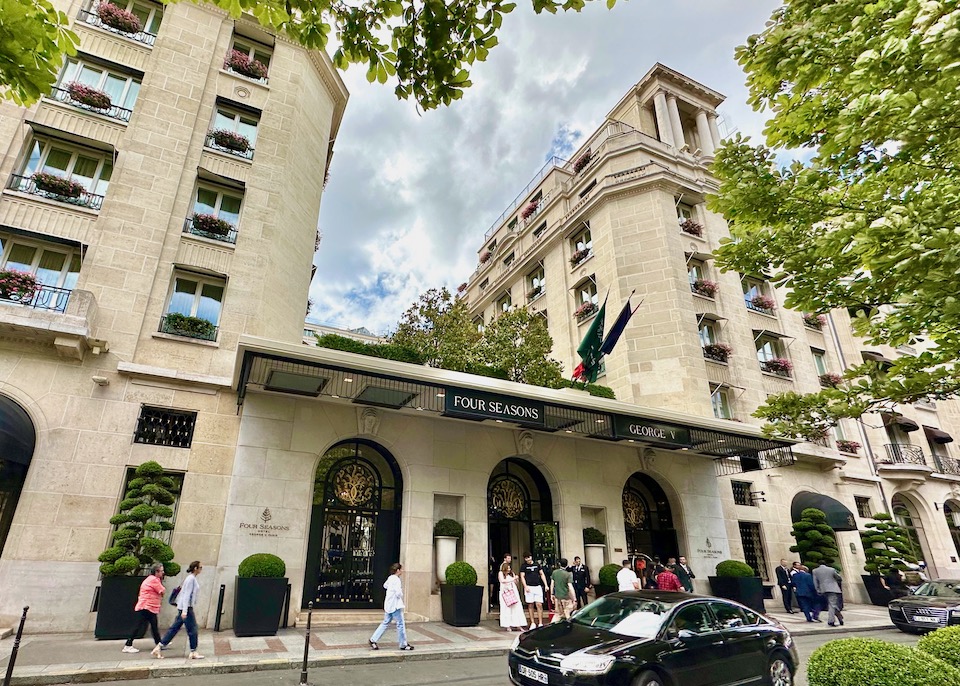 Exterior of the Four Seasons George V with two stone towers and wrought iron and gold doors on a tree-shaded street in Paris