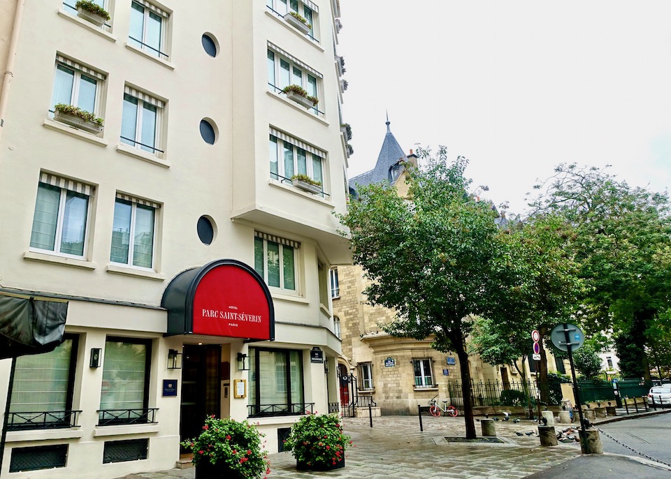 The modern exterior of the Parc Saint Severin hotel with a red awning and a row of little round windows on a tree-lined street in Paris