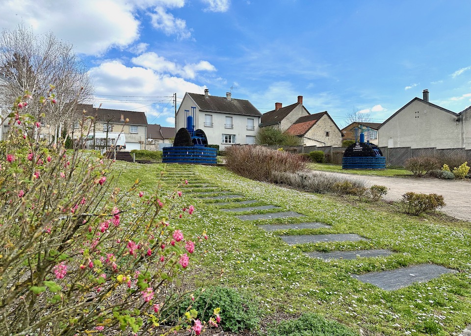 Green grass and pink budding flowers with a trail leading up to an old wine press painted blue with traditional houses in the French countryside village of Passy Grigny in the Champagne region of France