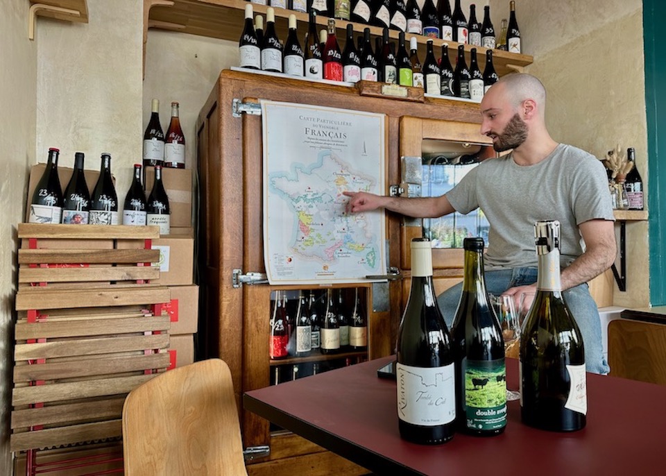 A sommelier points out French wine regions on a map with wine bottles on the table in front and shelves behind