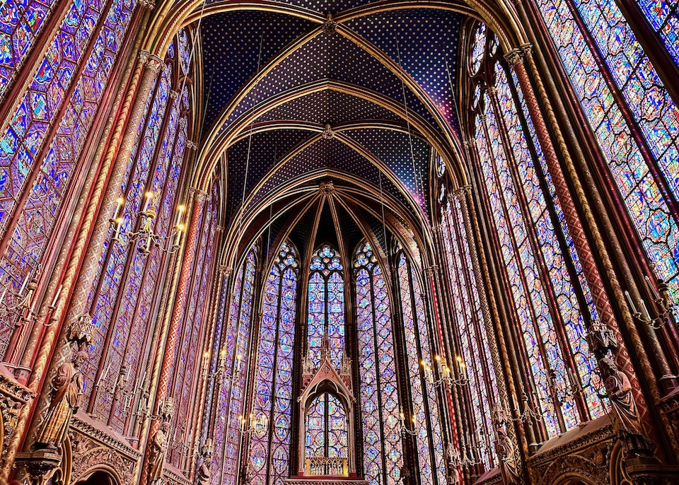 Multicolor stained glass windows encircling a chapel with gold columns and arches, and a vaulted ceiling painted in deep blue with stars inside Sainte-Chappele in the 1st Arrondissement of Paris