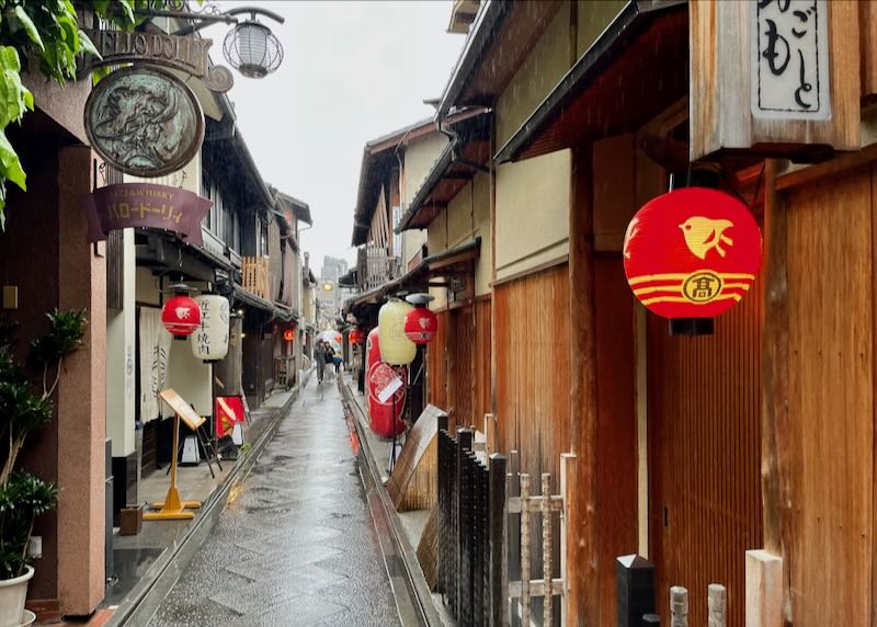 People walk through a rainy narrow alley lines with signs and red lanterns.