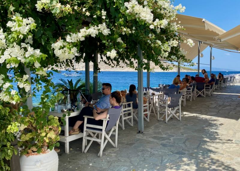 Diners at tables topped with sun umbrellas and bougainvillea, on a stone terrace overlooking the sea 