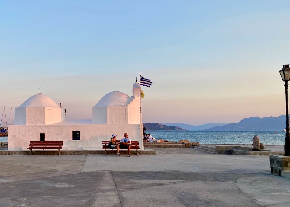 Two people sit on a bench in front of a small white greek chapel on the sea