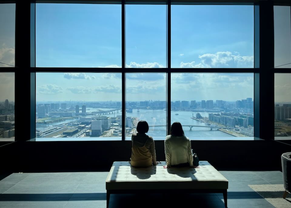 Two women sit on a bench in front of a large window with waterside views of Tokyo