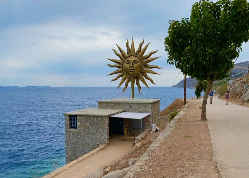 Boxy stone building perched on a cliff over the sea, topped by a large metal sun statue