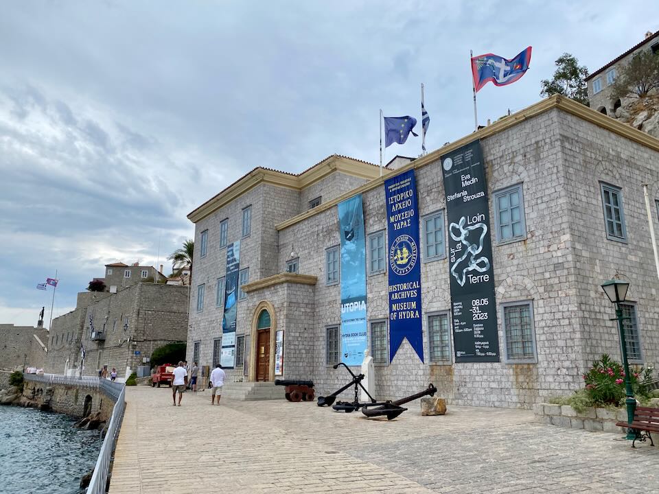 Greek flags fly on a stone building fronted with cannons along a stone harbor