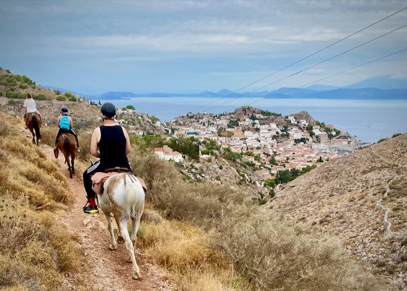 Three people riding horses along a trail overlooking a rustic Greek town and the sea
