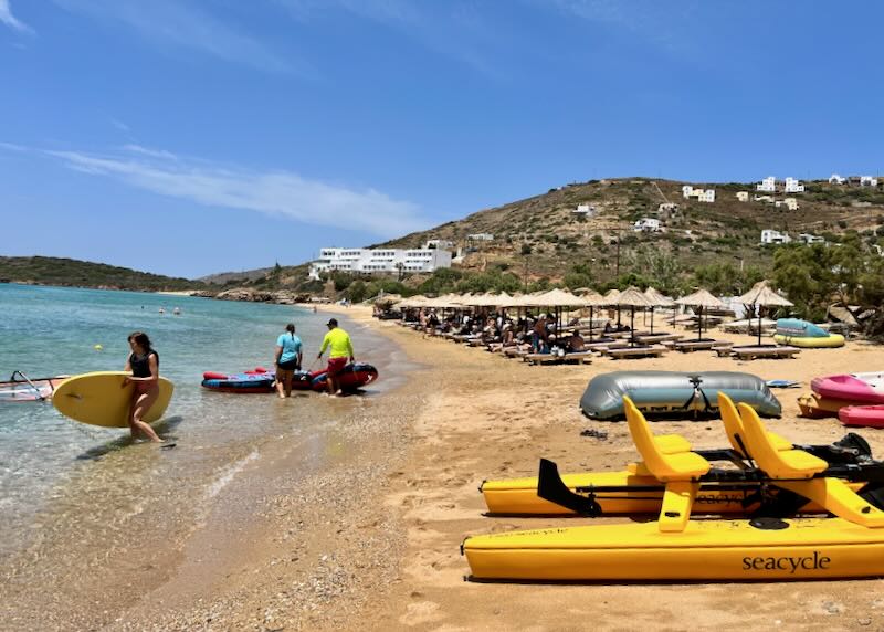 People on a golden sandy beach carry kayaks, paddleboards, and caramarans